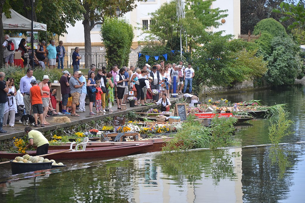 Mercado flotante de L'isle sur la sorgue con gente en la orilla del río y botes en el río llenos de frutas y verduras y vendedores vestidos con ropa provenzal