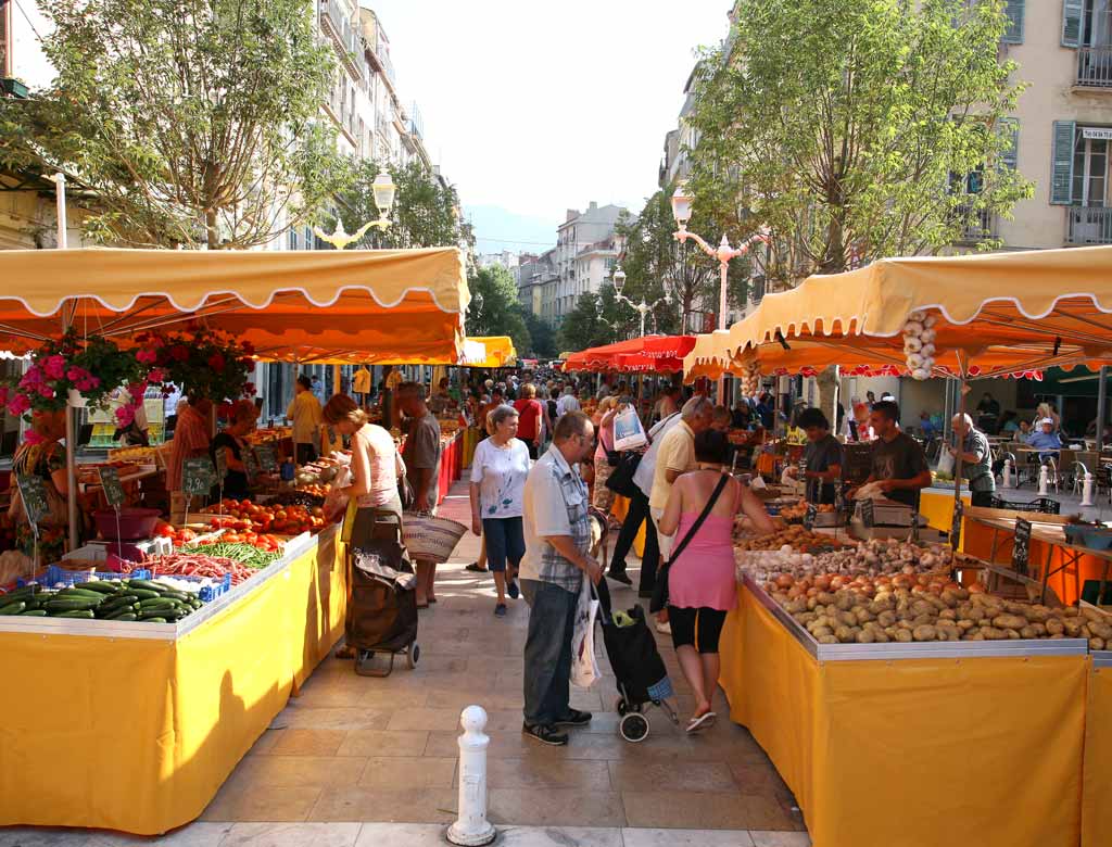 Mercado de Toulon Provence mirando filas de puestos uno frente al otro con manteles amarillos y sombrillas naranjas apiladas con frutas y verduras y gente en el medio