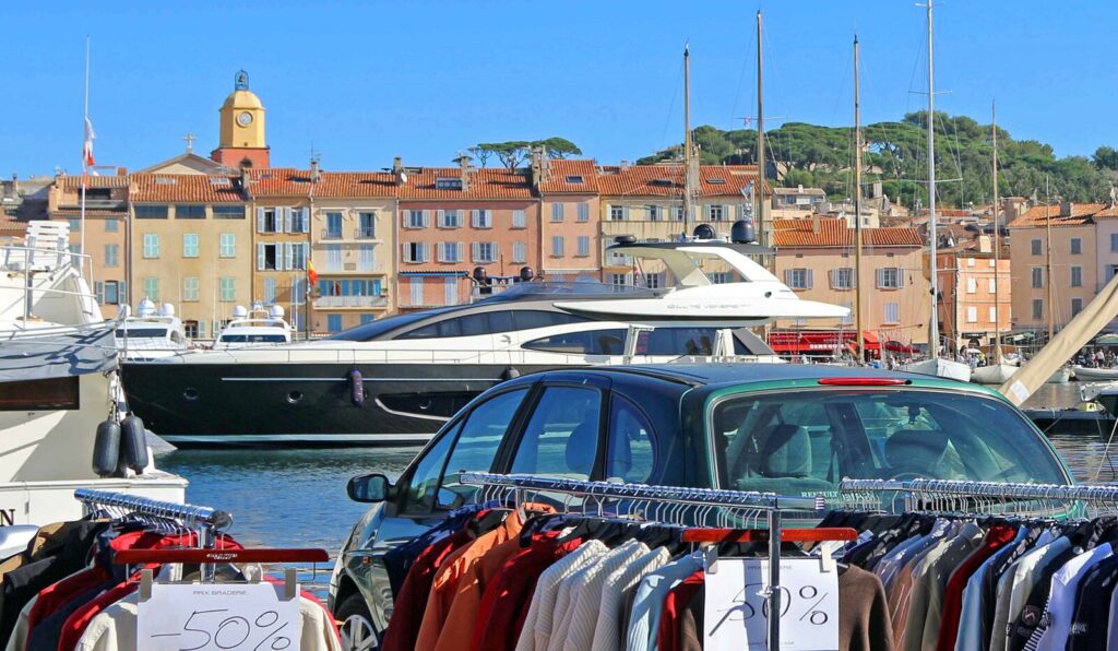 Grande Braderie de Saint Tropez con tiendas colocando percheros en el exterior. Este de la tienda que muestra percheros, un coche delante del agua y yates y casas detrás.