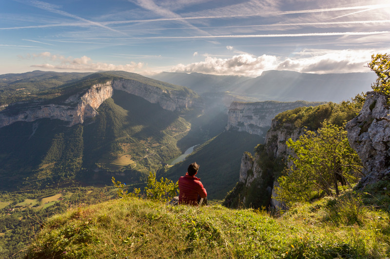 Caminante en Bercors mirando el desfiladero de Bourne muy por debajo con montañas onduladas en el fondo y un cielo azul con nubes y estelas de vapor