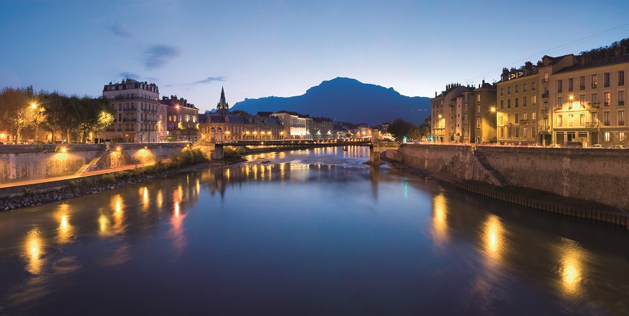 Grenoble al atardecer desde el río al frente con la ciudad en dos orillas visibles en el agua y una montaña detrás