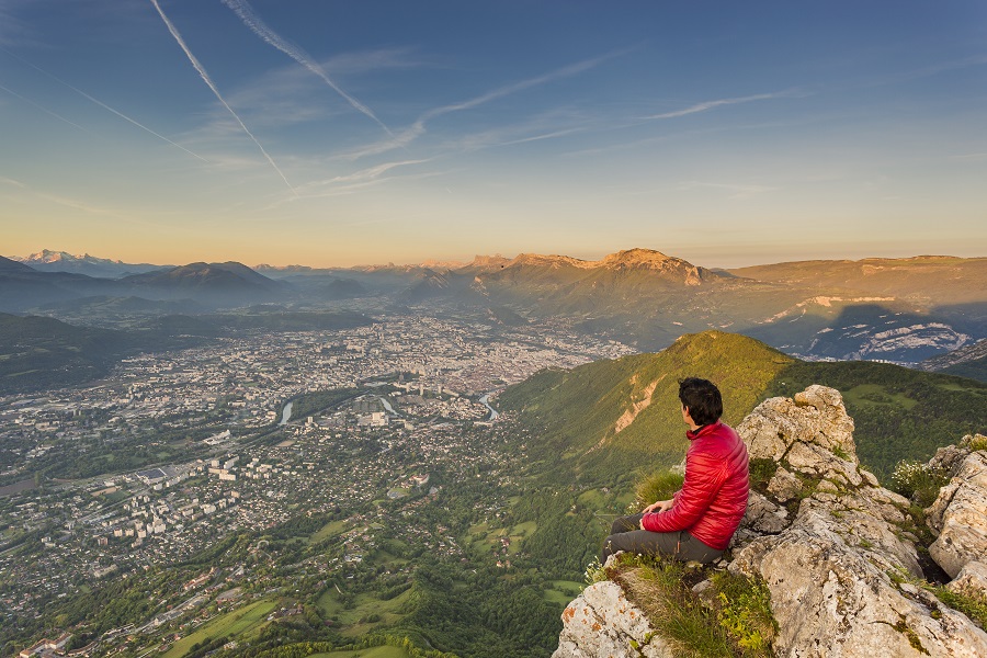 Una persona sentada en una roca alta mirando un largo camino hacia Grenoble con altas montañas y un cielo lejano