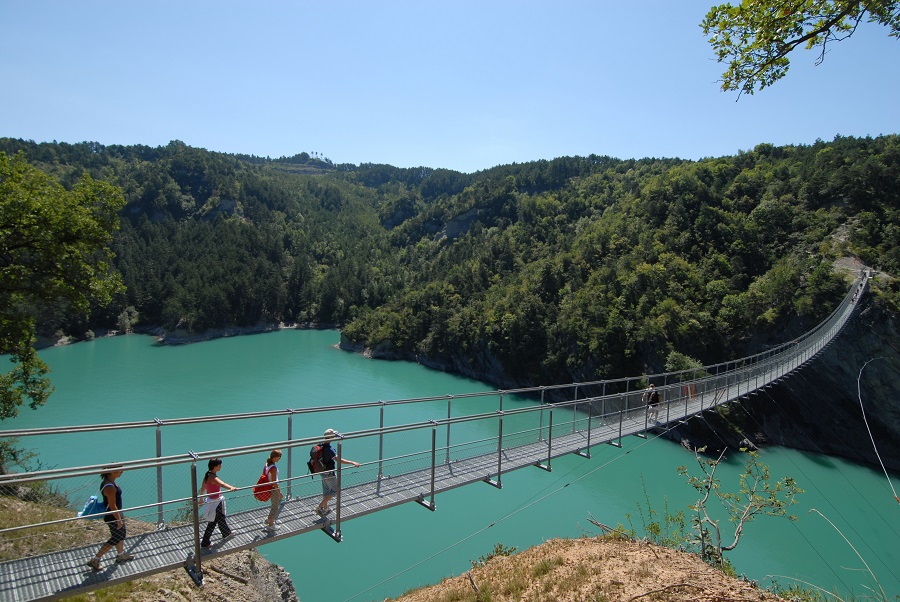 La gente caminando por un puente peatonal sobre lakd Monteynard y el lago debajo y en la distancia colinas cubiertas de árboles