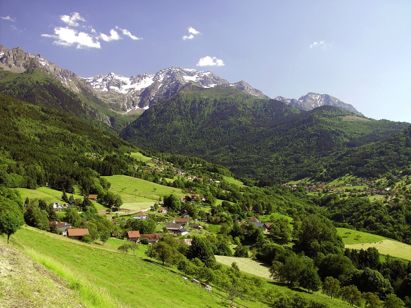 Balcons de Belledonne que muestra montañas cubiertas de nieve en el fondo, verdes colinas boscosas en el medio y ondulantes pastos verdes con edificios de granja rojos al frente.