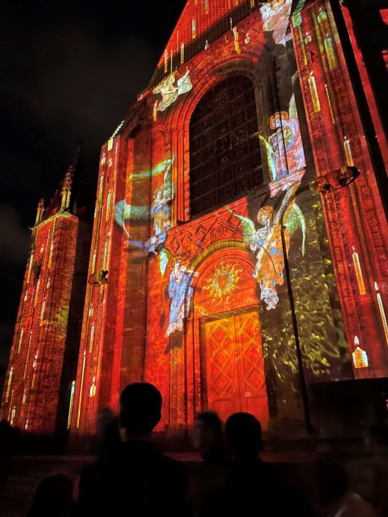 Catedral de Le Mans iluminada en rojo con ángeles azules y coloridos subiendo al frente