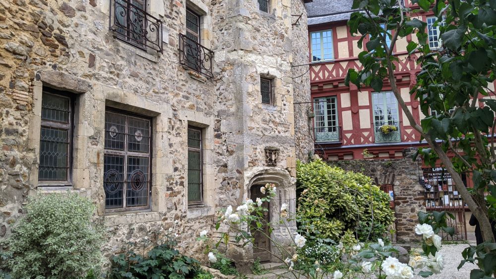 Bed and breakfast de Le Mans que muestra un gran edificio de piedra con un jardín de rosas blancas en la parte delantera y un edificio con entramado de madera en la parte trasera