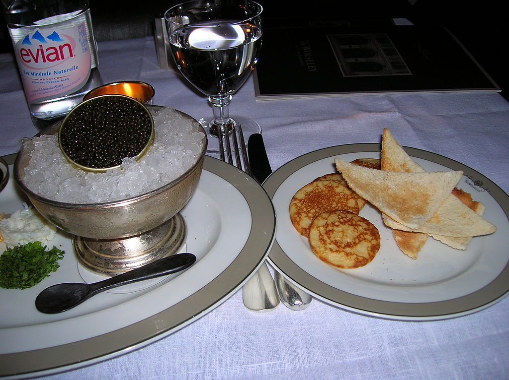 Caviar beluga en una olla pequeña con hielo en un plato de plata en un plato con una cuchara a la izquierda. Blinis y crema agria en el plato de la derecha