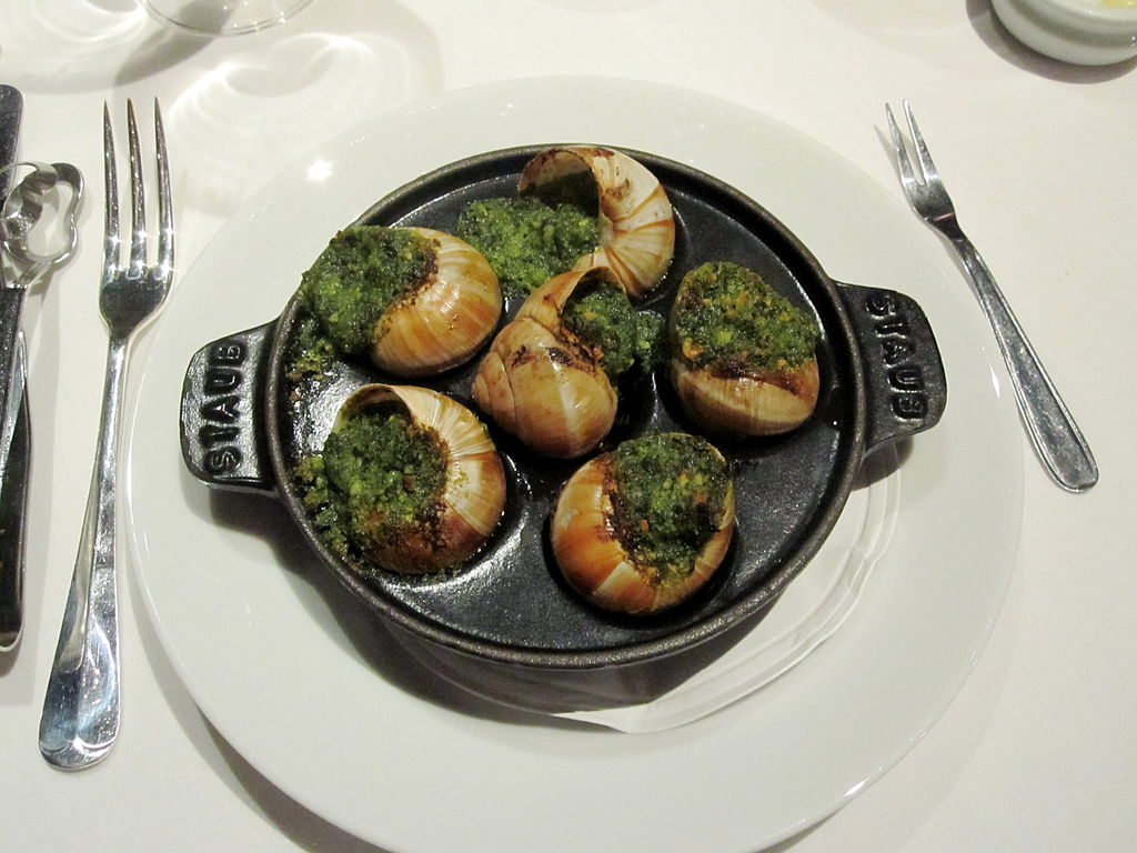 Un plato de hierro de forma especial de caracoles cocinados con perejil y ajo sobre un fondo blanco.