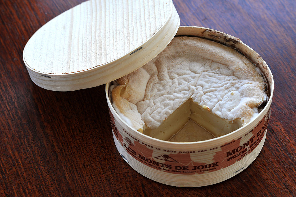 Una caja redonda de queso Vacherin Mont d'Or con una tapa equilibrada en un lado de la caja y una pieza cortada para revelar una parte superior corrugada y queso en el interior.