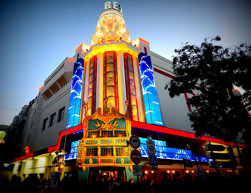 Cine Le Grand Rex en París. Frente que muestra un gran edificio antiguo con una gran fachada colonial y una cúpula en la esquina, todo iluminado con luces rojas que rodean ambas esquinas.