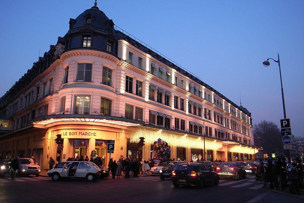 Los grandes almacenes de París Le Bon Marche en la noche del día de Navidad. Fachada iluminada en el edificio de la esquina, ventanas iluminadas y la multitud delante