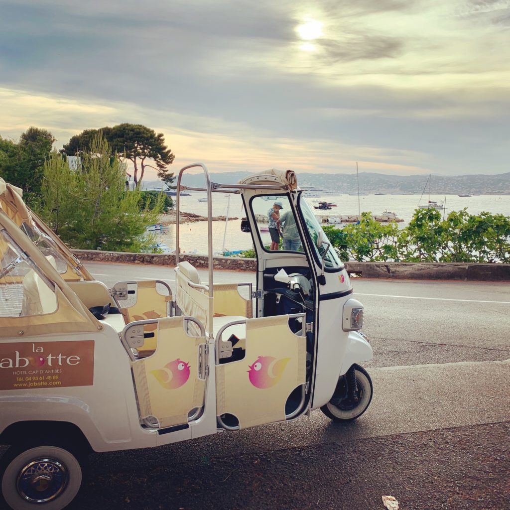 Little Tuk Tuk de La Jabotte estacionado en un camino con vista al hotel de mar con su nombre en el lugar estacionado 