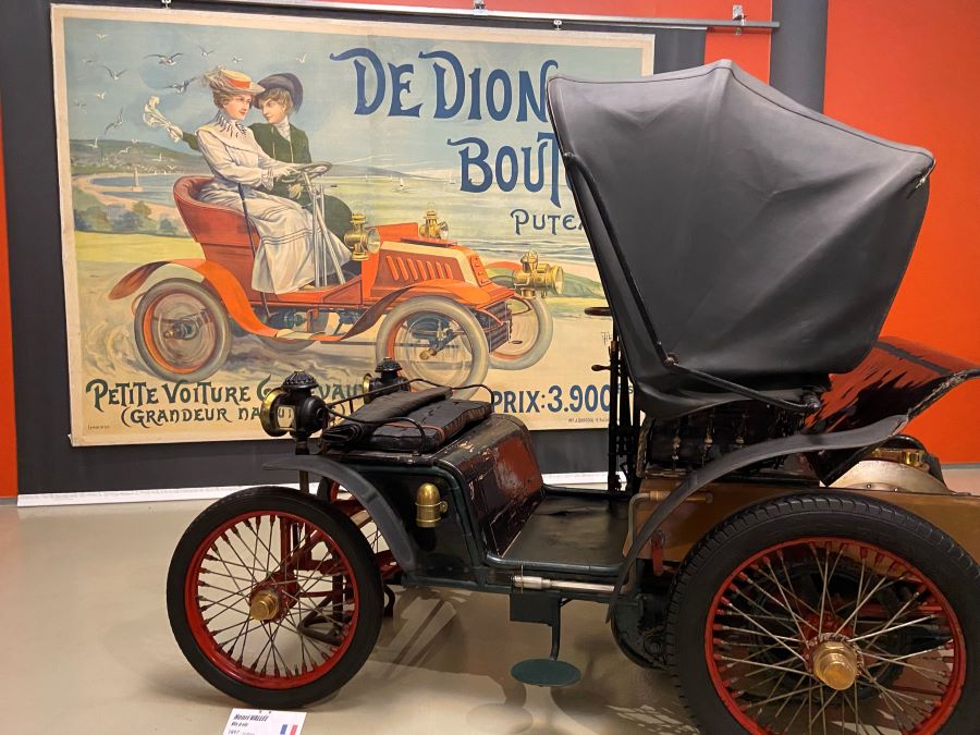 El auto encapuchado de De Dion Boulton frente a un póster que muestra a mujeres de principios del siglo XX en su fabuloso auto