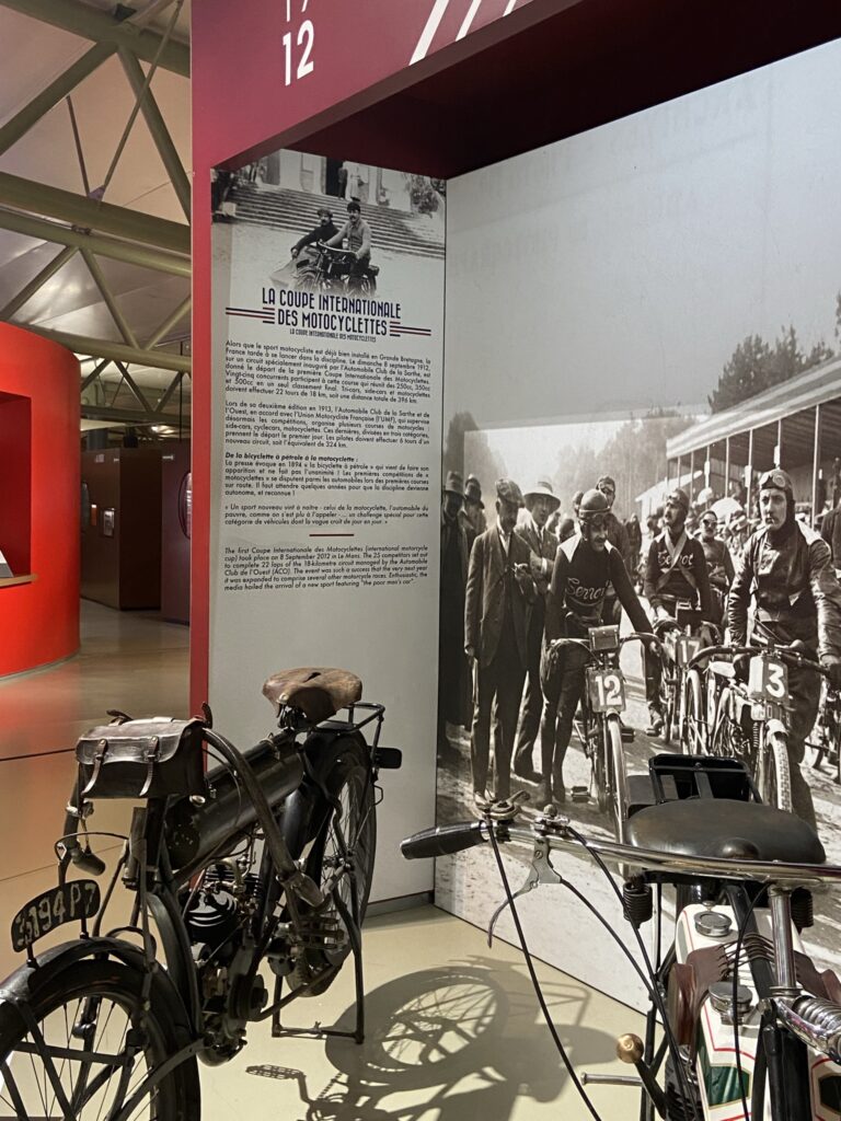 La sección de motocicletas del Museo de Le Mans con una bicicleta antigua en un soporte y un gran cartel con una explicación en la parte posterior.