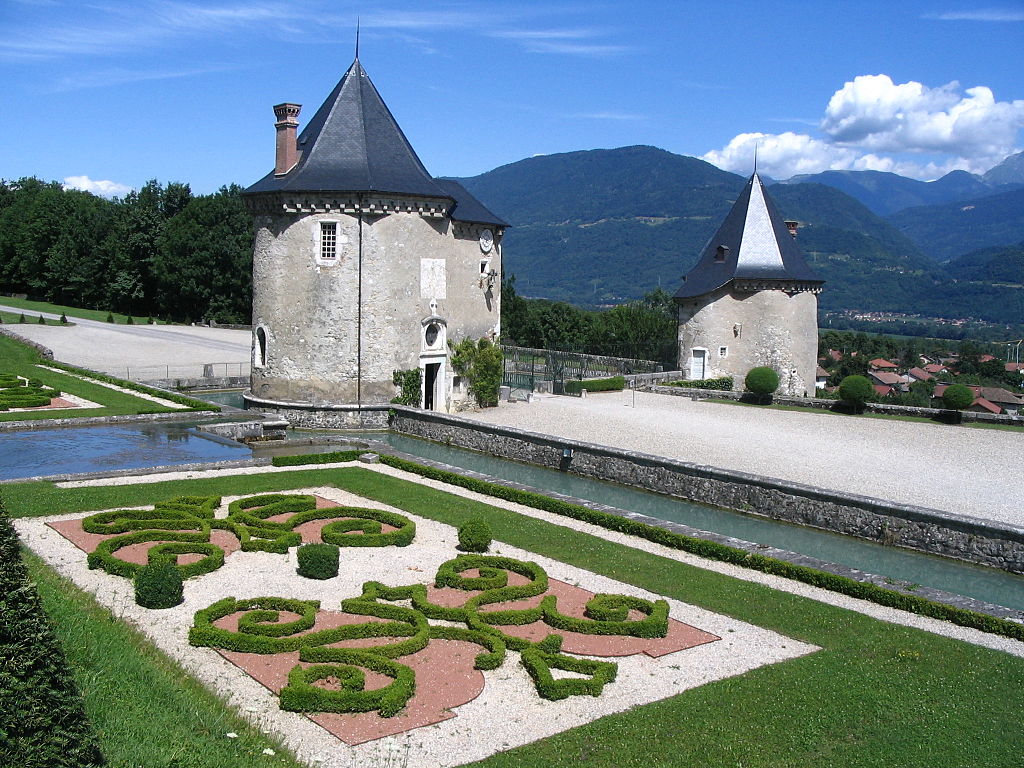 Jardines del Château du Touvet con un parterre formal de pequeños parterres de flores cercados en cajas en un rectángulo. Dos torres de piedra y un camino y una montaña detrás