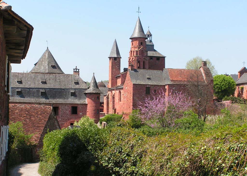 El pueblo de Collonges-la-Rouge mostrando arbustos delante con casas de piedra roja con torres y cimas cónicas detrás