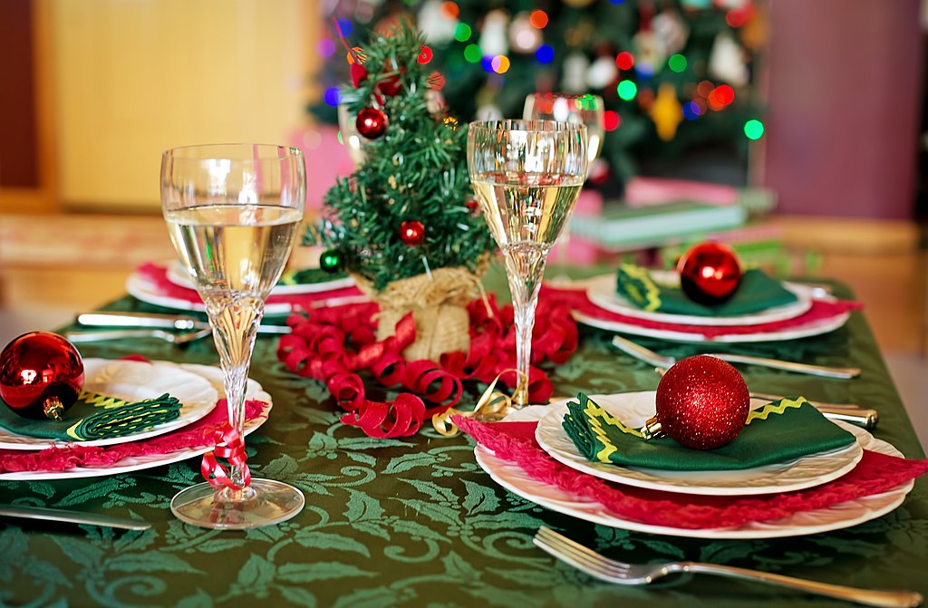 Mesas puestas para Navidad con un árbol decorado al fondo, y una mesa con mantel verde, adornos rojos y cintas y copas de champán