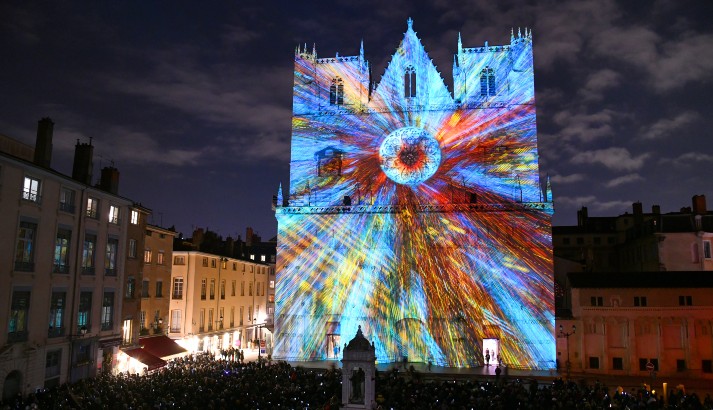 iluminación de la fachada de la Catedral de Lyon para un festival con luces en su mayoría tonos de azul y rojo que van desde un rosetón central por la noche con multitudes en el frente