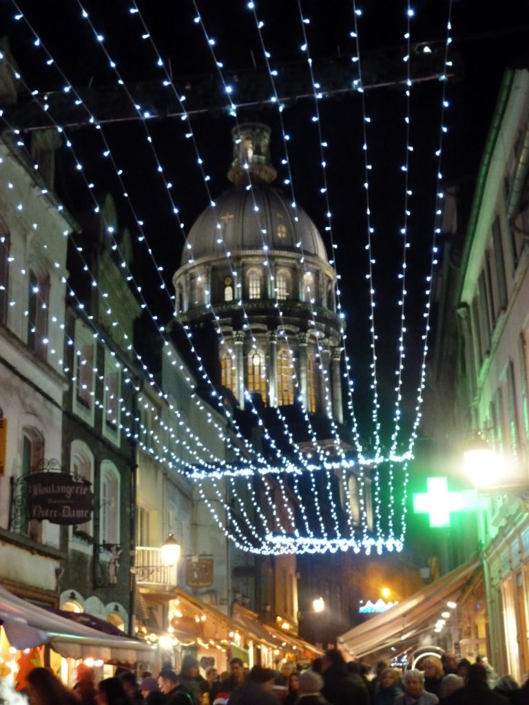 Vista a lo largo de la concurrida calle con luces de colores en el aire y la cúpula de la iglesia de Boulogne en el mercado navideño de fondo