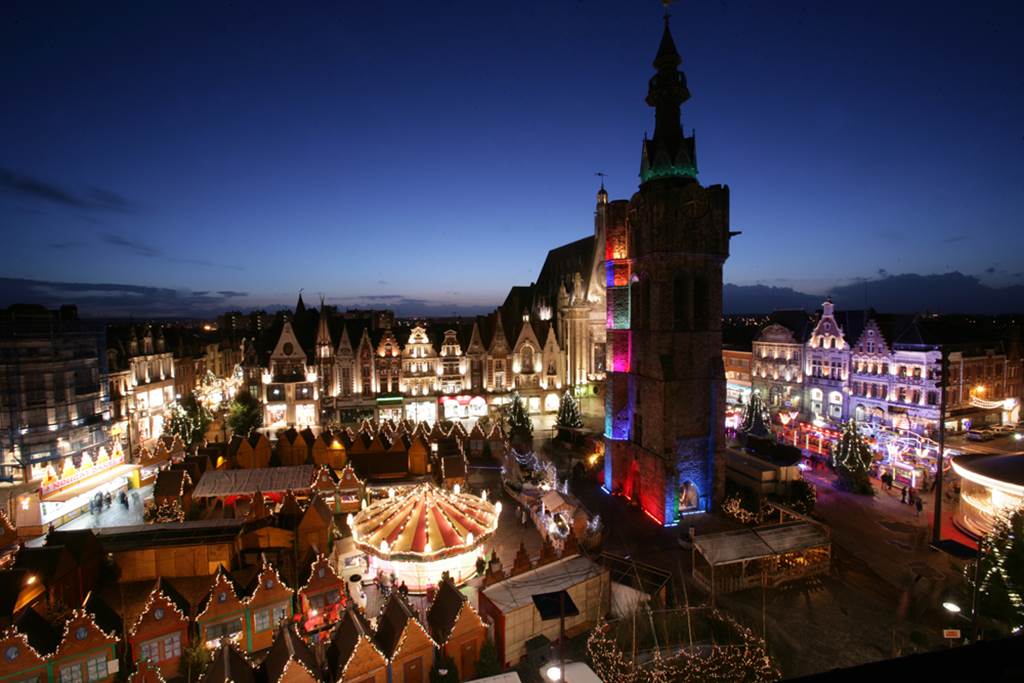 Vista aérea del mercado navideño de Bethune con campanario que se eleva sobre la plaza con carruseles y exhibiciones de bienes y personas 