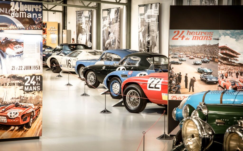 Coches antiguos estacionados en el museo de Le Mans desde atrás mostrando el lado izquierdo con números y por encima de grandes gradas con personajes famosos en ellos