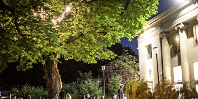 Noche de apertura del Festival de París que muestra un edificio clásico iluminado por la noche a la derecha, un árbol y un enorme césped iluminado con asistentes al festival sentados y de pie