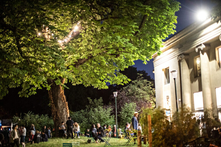 Noche de apertura del Festival de París que muestra un edificio clásico iluminado por la noche a la derecha, un árbol y un enorme césped iluminado con asistentes al festival sentados y de pie