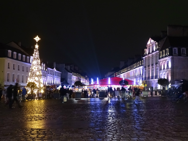 Un villancico por la noche con un árbol en llamas con una estrella en la parte superior izquierda, un grupo de velas iluminando el mercado con chozas en llamas detrás y edificios iluminados a la derecha