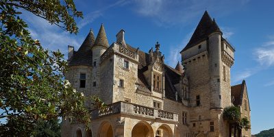 Chateau des milandes desde el frente que muestra una gran torre a la derecha, techos de piedra, paredes y balcones que dan a un camino de grava y árboles a la izquierda