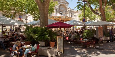 Vista exterior de un restaurante en la terraza de Avignon con mesas, sillas y sombrillas delante a la sombra de árboles y edificios antiguos detrás