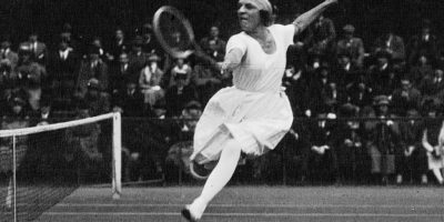 Suzanne Lenglen jugando tenis en 1920. Vieja foto en blanco y negro de ella en el aire haciendo un golpe de revés con un brazo levantado detrás de ella. ella esta en faldas largas