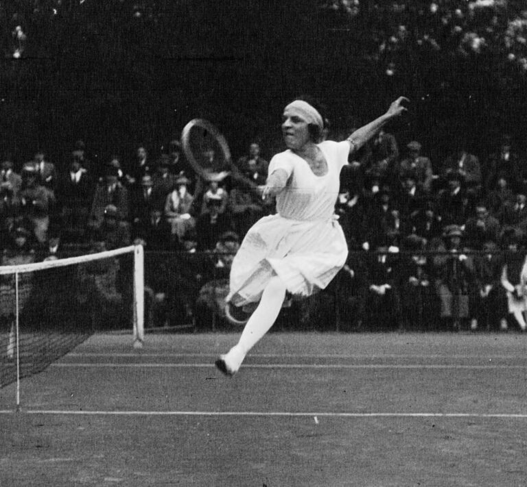 Suzanne Lenglen jugando tenis en 1920. Vieja foto en blanco y negro de ella en el aire haciendo un golpe de revés con un brazo levantado detrás de ella. ella esta en faldas largas