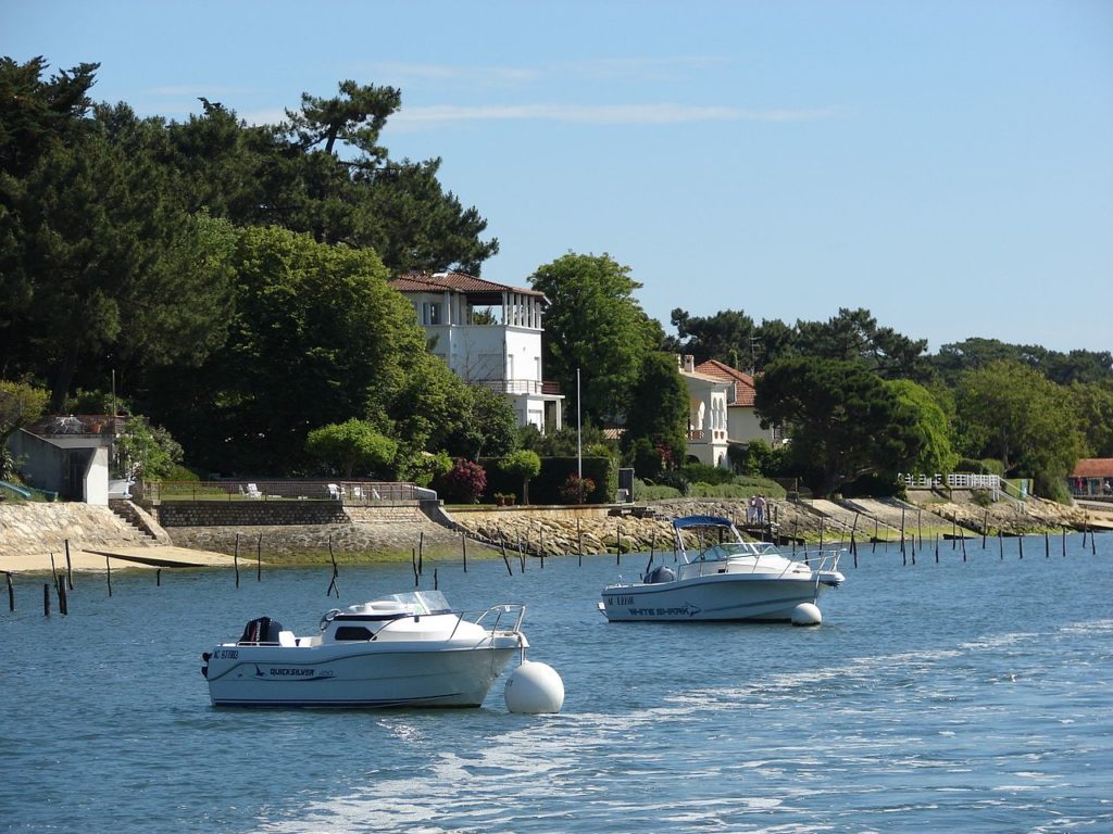 2 lanchas ancladas en el mar azul en la bahía de Archachon con paredes de piedra, villas y bosques al fondo