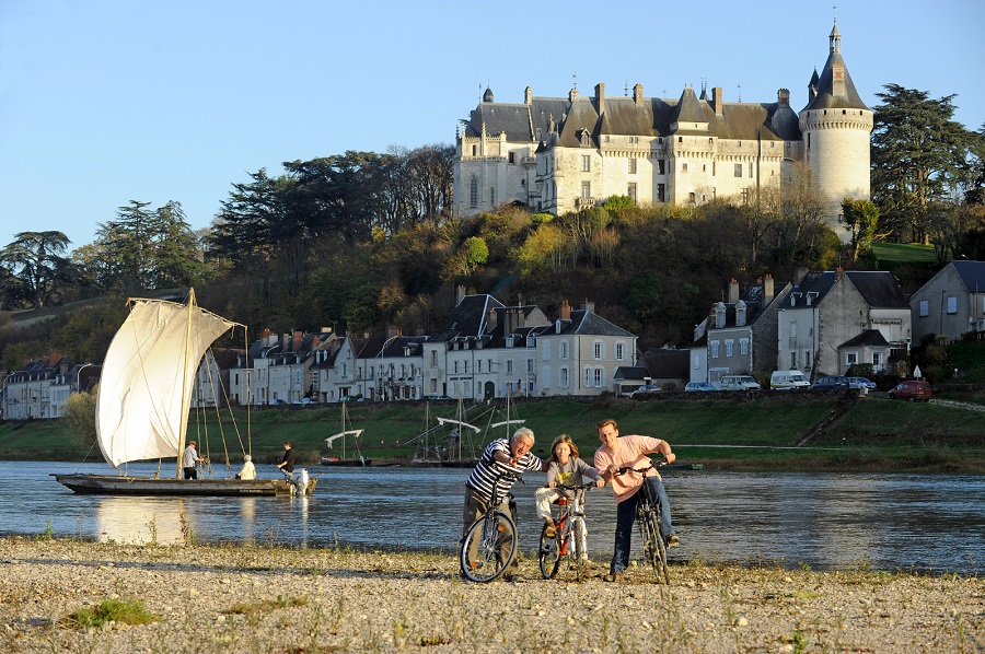 Tres ciclistas descansan junto al río Loira con Chaumont encaramado en la colina detrás. Están frente al fotógrafo y expresando algo a un niño. Un velero primitivo en el río