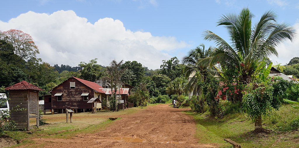 Una pista de tierra en Savall en la Guayana Francesa con tierra roja y casas a la izquierda con techos de hierro corrugado y árboles