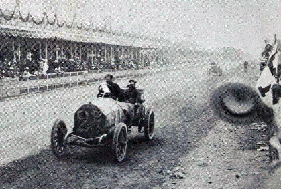 Nazzaro llegando a la carrera de Le Mans de 1906. Soporte con espectadores detrás; un coche con dos conductores en la pista de carreras