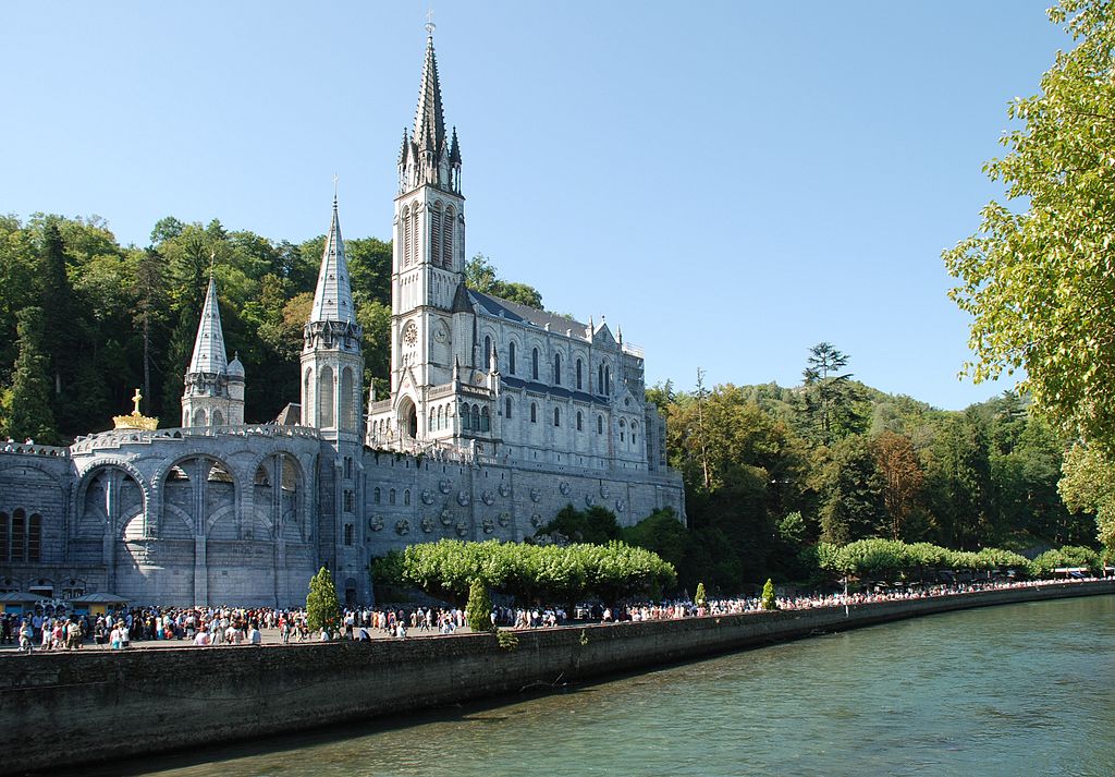 Basílica de Lourdes a orillas del río. El edificio del siglo XX tiene torres altas y muchos peregrinos en las orillas.