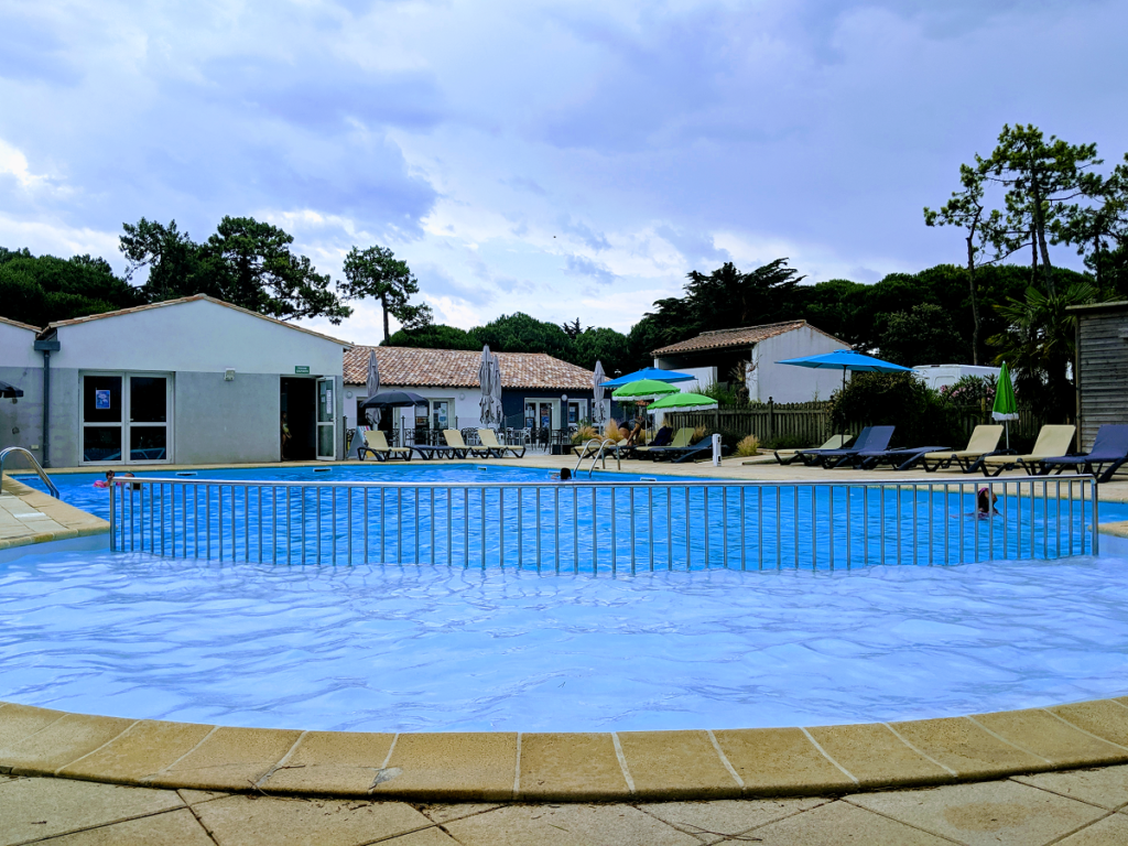 Vista del camping de la piscina al aire libre con un soiroit con una piscina azul en primer plano y una cabaña de un solo piso en la parte posterior