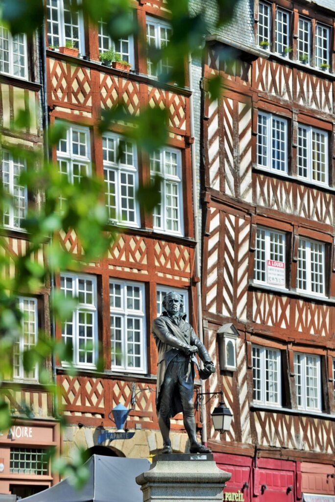 Dos plantas superiores de casas con entramado de madera en Rennes. De color rojo y blanco y marrón y blanco con un árbol al frente y una estatua