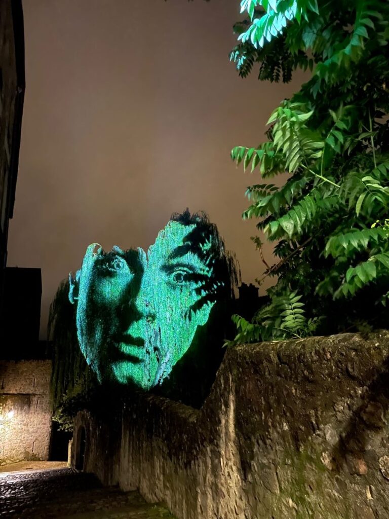 La iluminación de Le Mans de noche y un enorme rostro colgado sobre un muro de piedra en verde