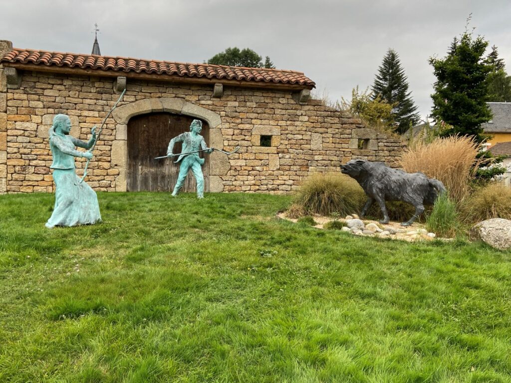 Un antiguo granero con puertas cerradas frente a un parche verde con dos figuras de bronce y horcas sosteniendo una bete du gevaudon a raya