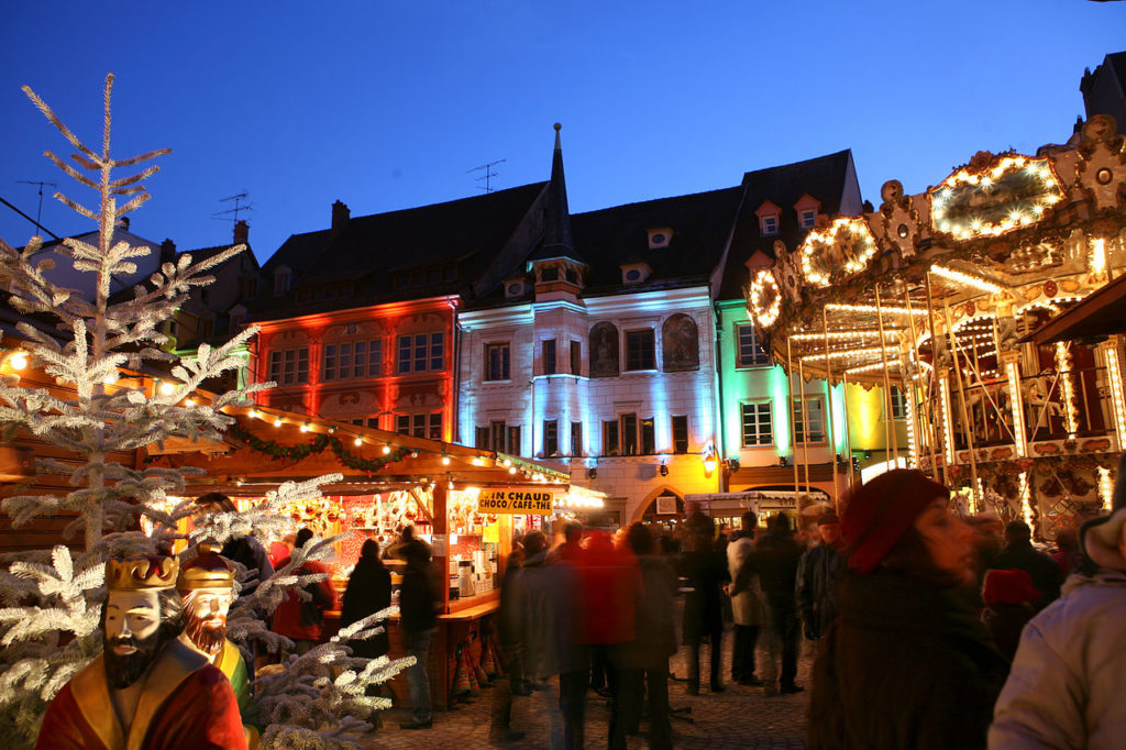 Mercado navideño de Mulhouse con edificios iluminados detrás, puestos y gente por la noche