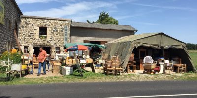 Brocante de Charreyrot en Auvernia. Vista exterior al otro lado de la carretera con un granero y una carpa grande y muebles de campo viejos derramándose sobre la hierba y un hombre caminando hacia la izquierda