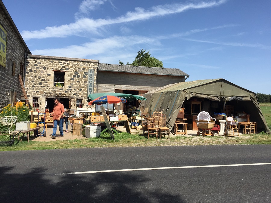 Brocante de Charreyrot en Auvernia. Vista exterior al otro lado de la carretera con un granero y una carpa grande y muebles de campo viejos derramándose sobre la hierba y un hombre caminando hacia la izquierda