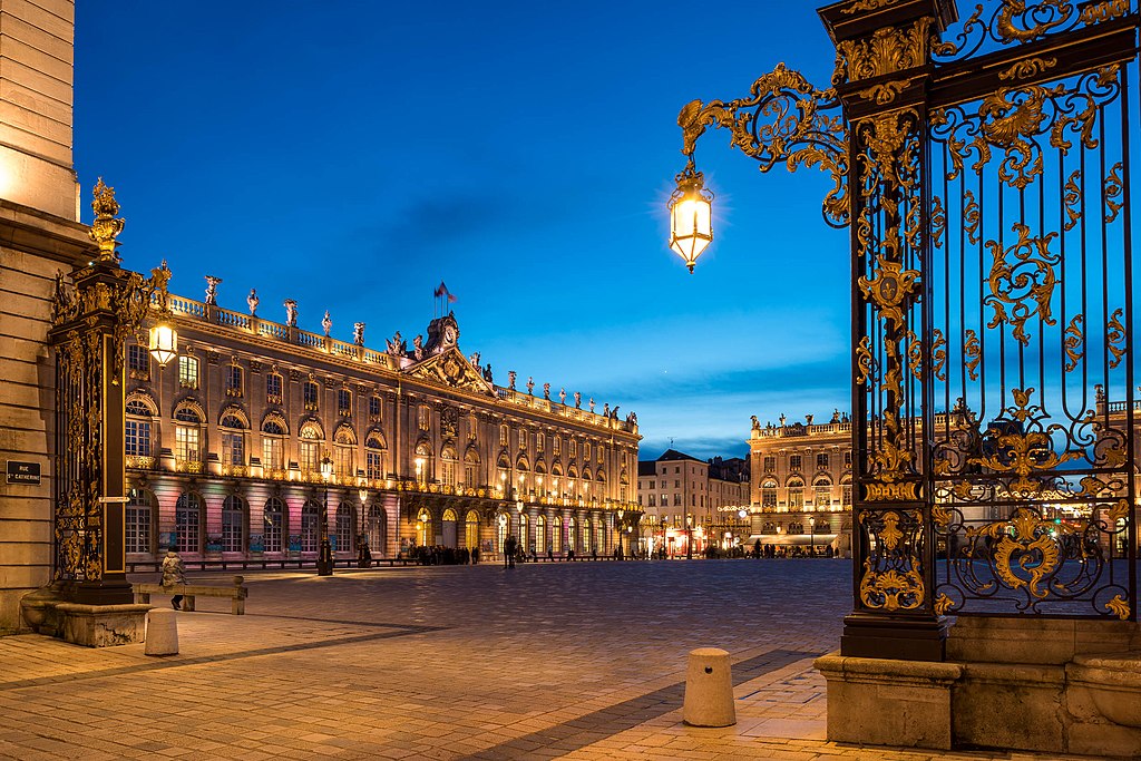 Place Stanislas en Nancy de noche que muestra una vista a través de una elaborada parrilla de hierro fundido en negro y dorado y un largo edificio clásico con focos