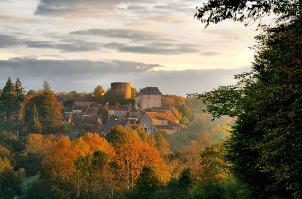 El pueblo de Saint-Sauveur en otoño con pinos verdes y árboles de colores otoñales en primer plano y un pueblo con un campanario y casas antiguas en el fondo